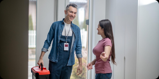 Uśmiechnięty technik w roboczym kombinezonie i koszuli, z identyfikatorem na szyi, wchodzi do domu, trzymając czerwoną skrzynkę narzędziową. Wita się z kobietą w różowej koszulce, która wskazuje mu kierunek wewnątrz domu. W tle widać drzwi wejściowe i część podwórka.