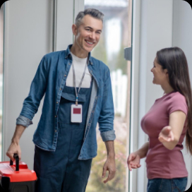 Uśmiechnięty technik w roboczym kombinezonie i koszuli, z identyfikatorem na szyi, wchodzi do domu, trzymając czerwoną skrzynkę narzędziową. Wita się z kobietą w różowej koszulce, która wskazuje mu kierunek wewnątrz domu. W tle widać drzwi wejściowe i część podwórka.
