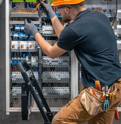 Elektryk w pomarańczowym kasku ochronnym i rękawicach roboczych pracuje przy dużej szafie elektrycznej, regulując przewody za pomocą śrubokręta. Stoi na małej drabince, a wokół pasa ma narzędzia, takie jak szczypce i śrubokręty, w pasie narzędziowym.